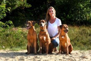 Iva zit gehurkt met drie van haar Rhodesian Ridgebacks op het zand